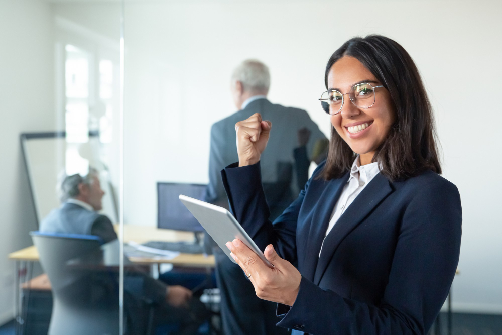 happy-female-professional-glasses-suit-holding-tablet-making-winner-gesture-while-two-businessmen-working-glass-wall-copy-space-communication-concept