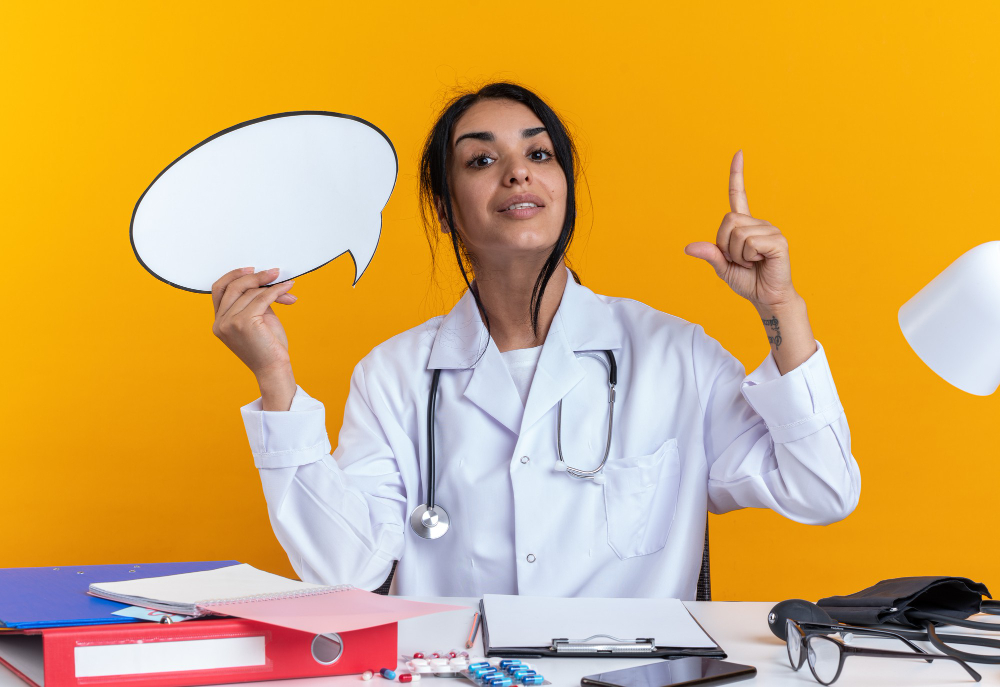 impressed-young-female-doctor-wearing-medical-robe-with-stethoscope-sits-table-with-medical-tools-holding-speech-bubble-points-up-isolated-yellow-wall