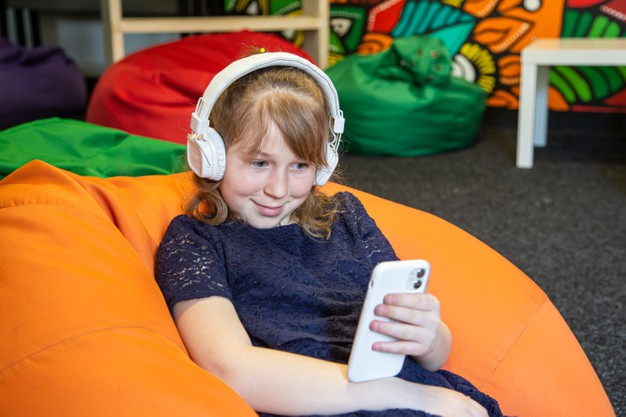 little-girl-uses-phone-listens-music-with-headphones-while-sitting-bag-chair_169016-11839