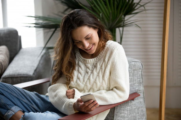 smiling-happy-millennial-woman-holding-smartphone-relaxing-armchair_1163-4851