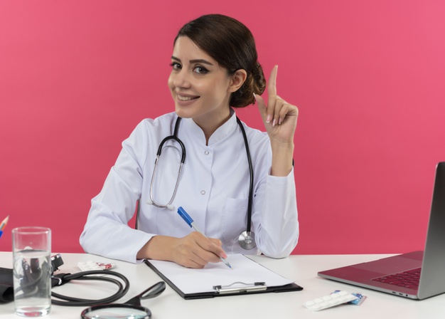 smiling-young-female-doctor-wearing-medical-robe-stethoscope-sitting-desk-with-medical-tools-laptop-holding-pen-raising-finger-isolated-pink-wall_141793-59395