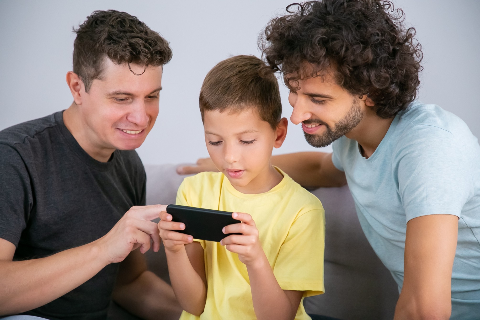 two-happy-dads-teaching-son-use-online-app-cell-boy-playing-game-mobile-phone-family-home-communication-concept (1) (1)