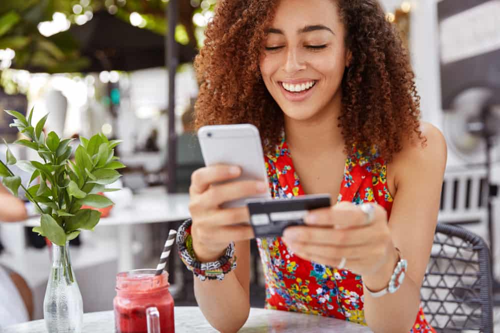 beautiful-dark-skinned-young-female-with-cheerful-expression-holds-smart-phone-credit-card-banks-online-makes-shopping-while-sits-against-cafe-interior