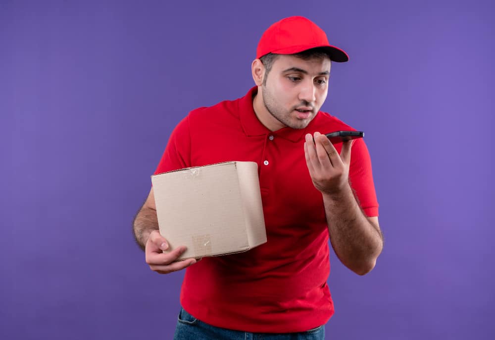 young-delivery-man-red-uniform-cap-holding-box-package-sending-voice-message-using-his-smartphone-standing-purple-wall